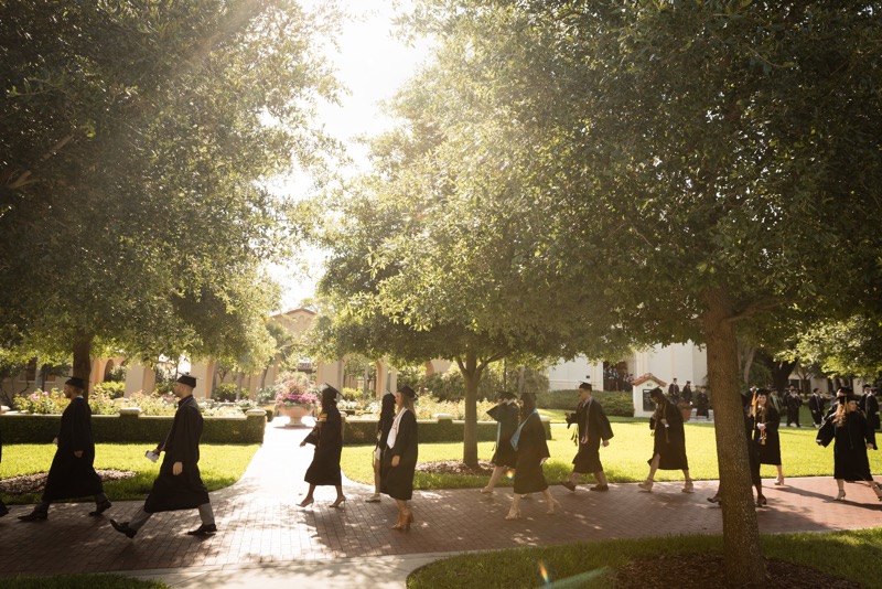 Commencement procession of students in caps & gowns through the Rollins campus, under large trees with the sun glowing through