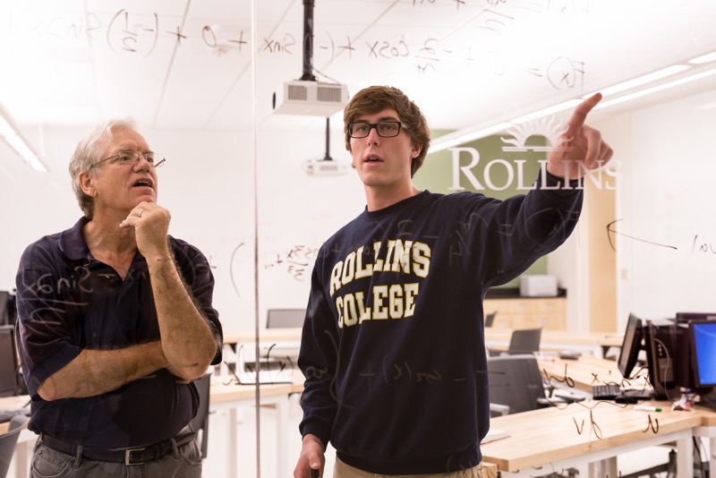 Professor has hand on chin, looking at a window wall of a Rollins science lab, standing next to a student discussing and pointing at a formula on the window.