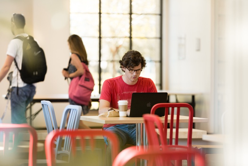 Male student at table in the Rollins Cafe, on his laptop with a coffee. Two students with backpacks walk past him.