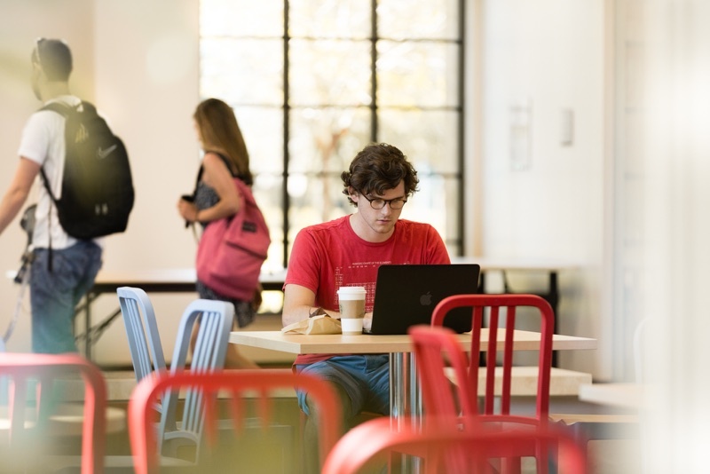 Male student at a table at a Rollins cafe, in front of his laptop as students walk by him on their way to class.