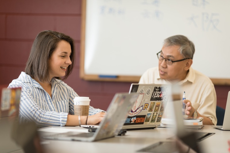 Professor and student meet to discuss a project. Student is smiling taking notes on a laptop as professor is talking.