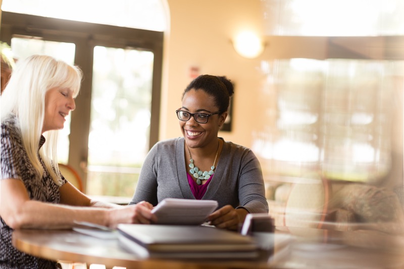 Professor and student meet to discuss a project. Student smiles and holds a notebook as professor explains the situation.